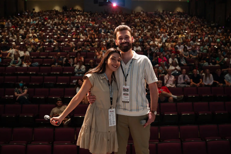 Two filmmakers stand on a stage with microphones with the seated audience in the theater in the background.