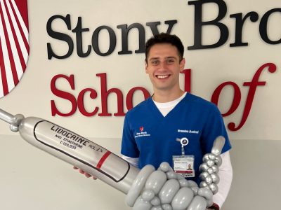 Axelrod holds a large balloon model of a lidocaine injection. He is wearing scrubs and standing in front of a Stony Brook School of Dental Medicine sign.